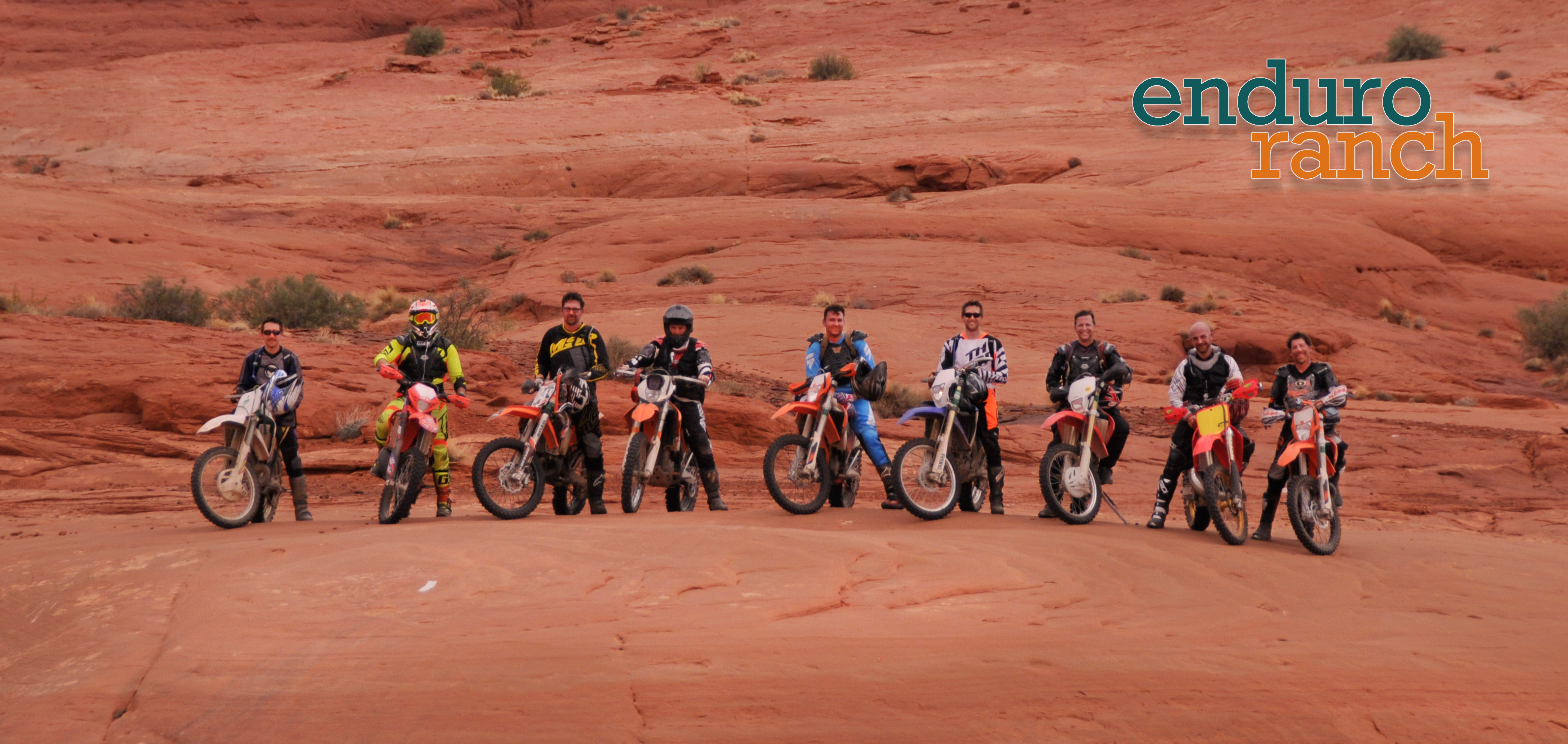 Shot of 9 dirtbike riders lined up on domed rock far away from camera.