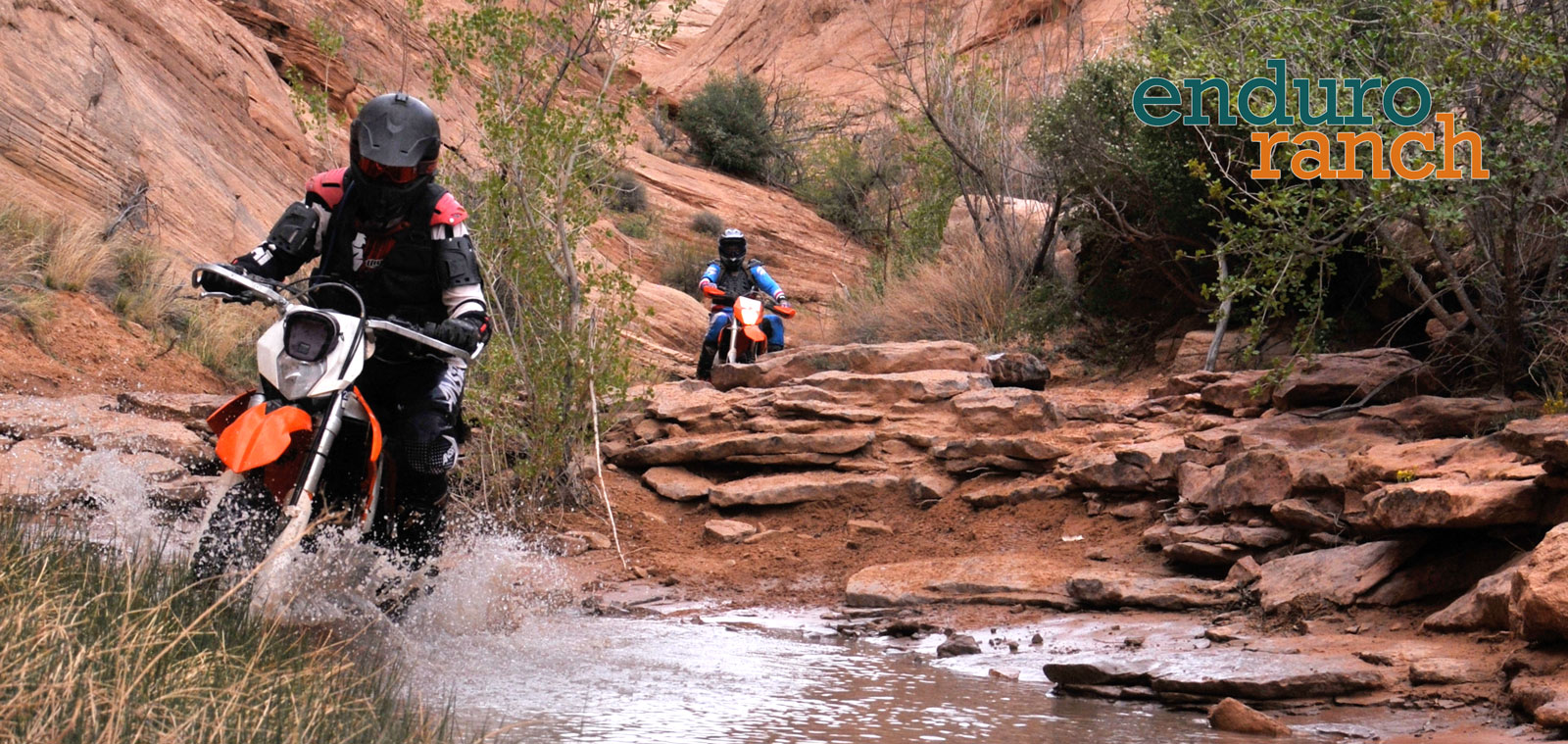 Two off road motorcycle riders in a small canyon bottom riding over rocks and through a creek.