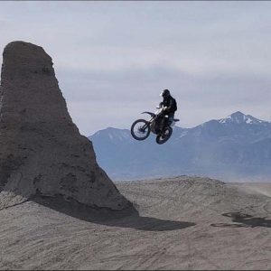 Dirtbiker catches air by small monument of barren dirt with snowcap mountains in the background.